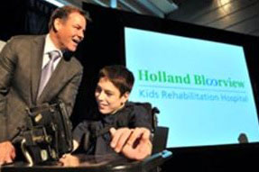 An adult standing on the left, and a child sitting on a wheelchair on the right, in front of a big screen showing the Holland Bloorview new logo