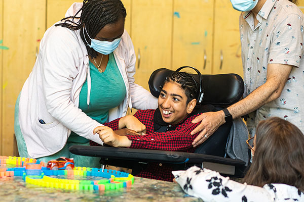 An adult on the left is helping a teen on a wheelchair with a hearing machine on. Another adult standing to the right.
