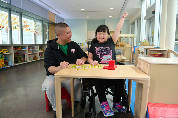 An adult sitting to the right of a child on wheelchair, playing a game