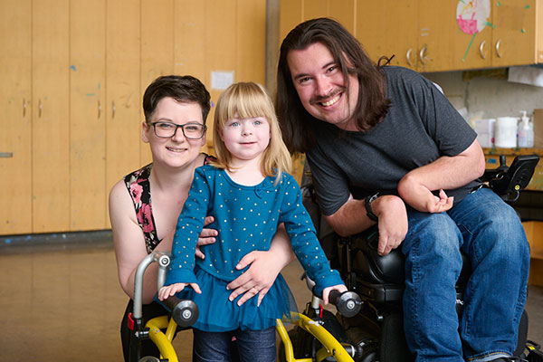 An adult is holding a child in the middle with a walker and to their right there is another adult, all with smiling face