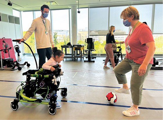 An adult kicking a ball, and another adult standing behind a child with a walker.