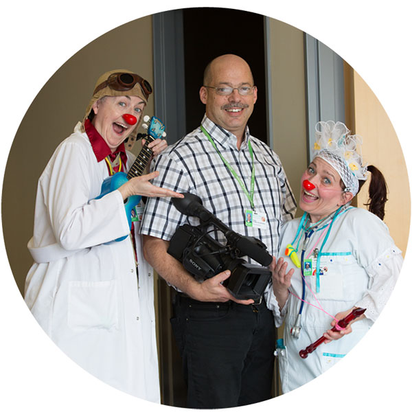 Three adults standing, the left and right are clowns while the middle one is holding a video camera