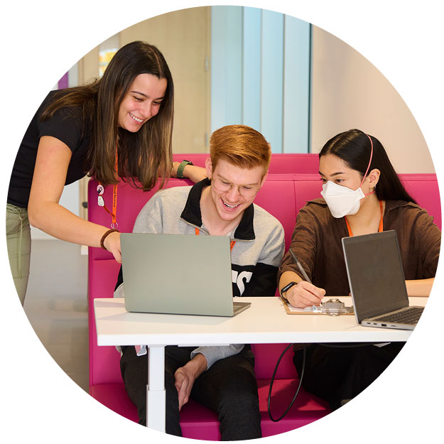 Three adults sitting to discuss something in front of some laptop computers. One adult is with a mask on.