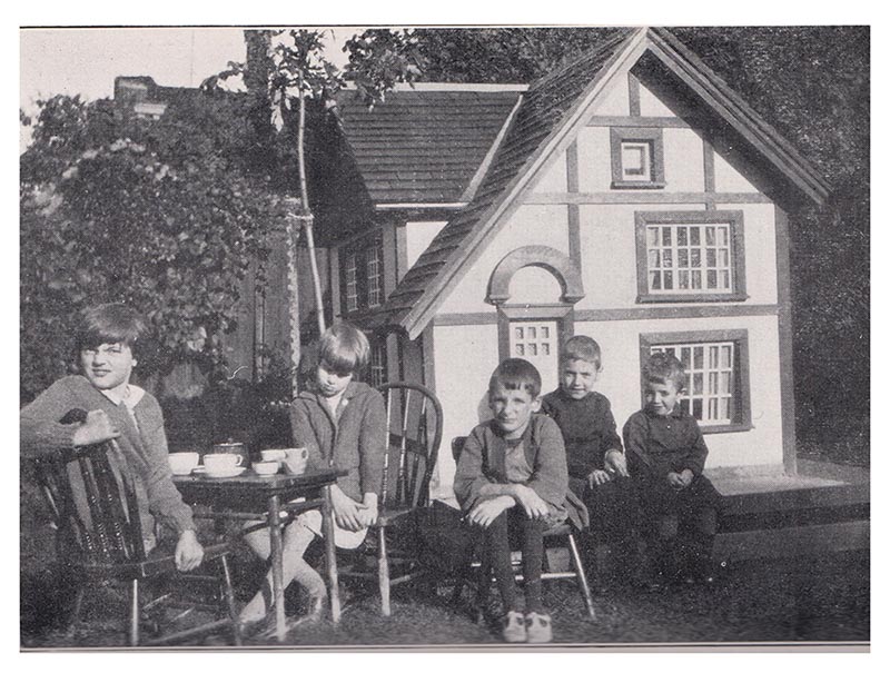 A back and white photo with some children sitting in front of a giant doll house.